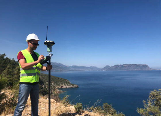 Geodetic engineer surveyor in white hard hat doing measurements with GNSS satellite receiver at the hill top above the sea
